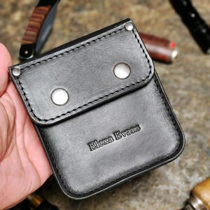 May include: Black leather wallet with the embossed text "Elexa Evans". The wallet has a flap secured by two silver-coloured press studs. The edges are stitched with black thread. Other tools and a knife are in the background.