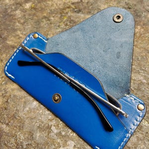 May include: A blue leather glasses case with white stitching and a snap closure. The case holds a pair of black-framed glasses with silver arms. The case is open, revealing the glasses inside.