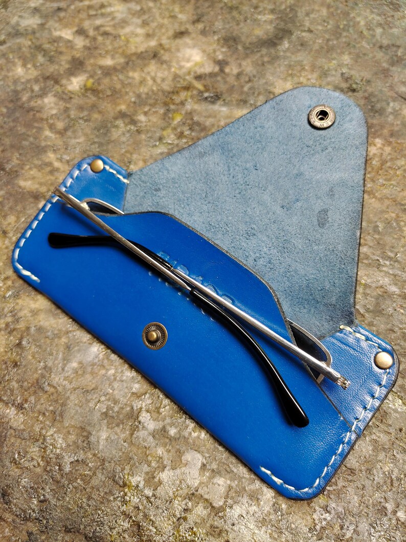 May include: A blue leather glasses case with white stitching and a snap closure. The case holds a pair of black-framed glasses with silver arms. The case is open, revealing the glasses inside.