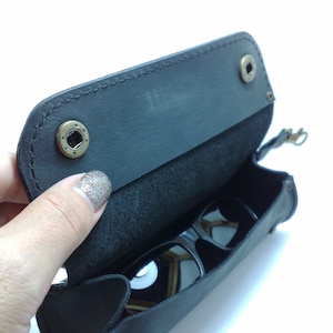 May include: A black leather sunglasses case with a flap that opens to reveal a pair of sunglasses inside.