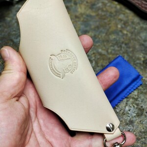Puede incluir: Funda de cuchillo de cuero beige con cierre y remaches met&aacute;licos. La funda tiene un logotipo circular en relieve y un accesorio para llavero. Un pa&ntilde;o azul es visible en el fondo.
