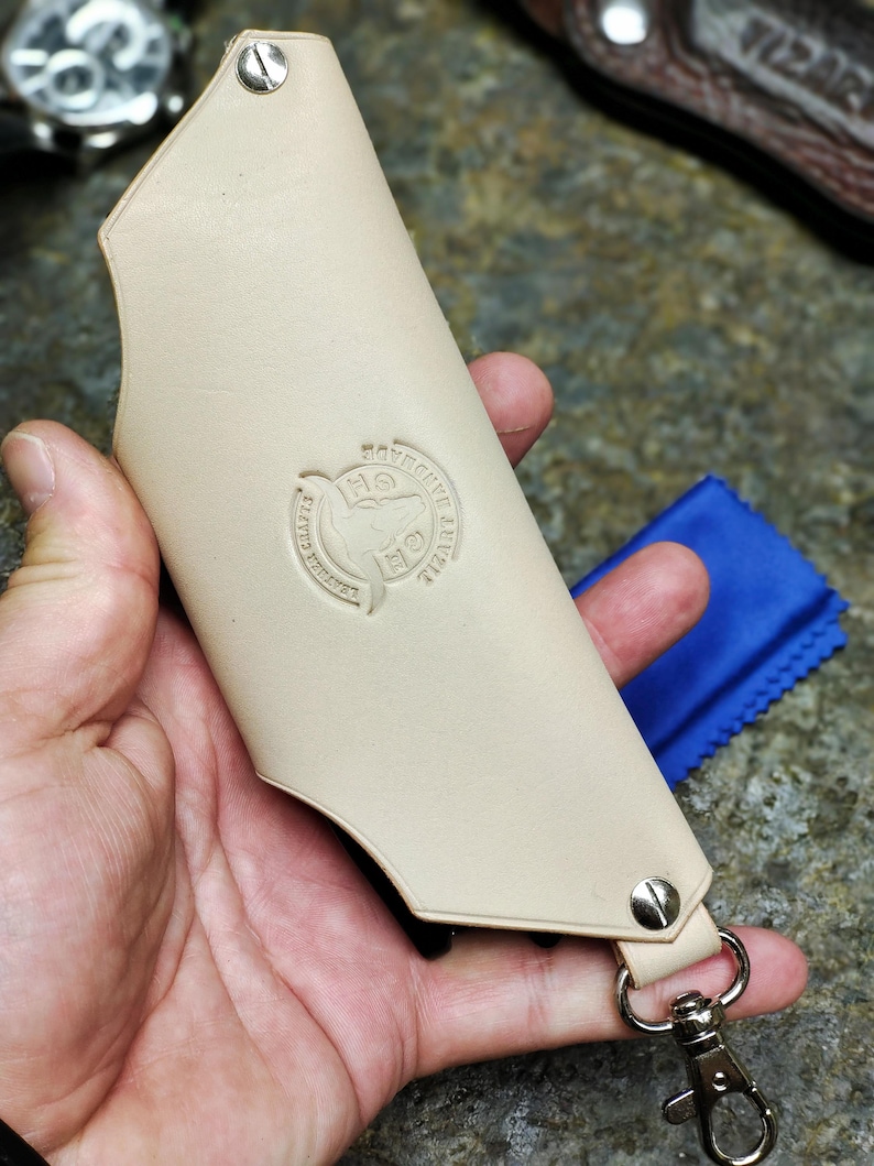 Puede incluir: Funda de cuchillo de cuero beige con cierre y remaches met&aacute;licos. La funda tiene un logotipo circular en relieve y un accesorio para llavero. Un pa&ntilde;o azul es visible en el fondo.