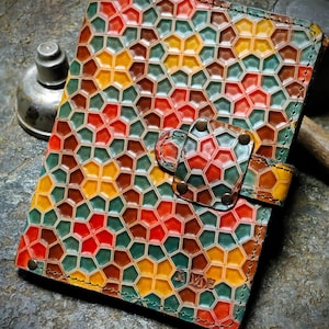 May include: A leather notebook with a geometric pattern in shades of red, green, yellow, and brown. The notebook has a flap closure with a metal button and the initials "R.M.E." embossed on the front.