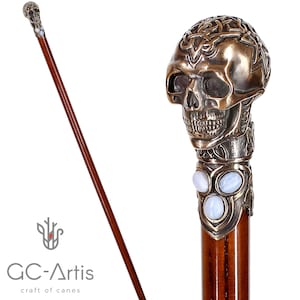May include: A brown walking cane with a skull-shaped handle. The handle is bronze-coloured with intricate detailing. The shaft is a polished brown wood. The cane features three oval-shaped, light blue stones. The text "GC-Artis craft of canes" is visible.