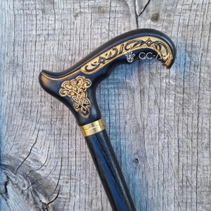 Close-up photo of the Celtic cane against weathered wood. The engraved handle with gold detailing stands out sharply, and the black shaft continues downward with a smooth, polished finish. The natural backdrop highlights the fine carving and elegant shape, giving the cane a strong, classic presence.