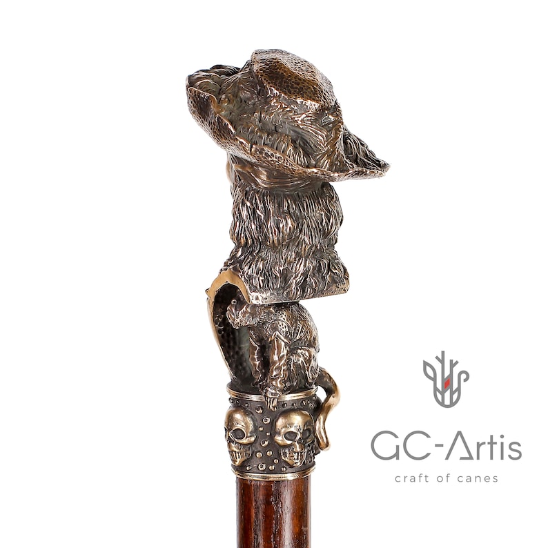 May include: A bronze walking stick with a detailed sculpture of a dog wearing a hat. The stick has a wooden shaft and a decorative skull design at the base. The sculpture is attached to the shaft with a metal ring.