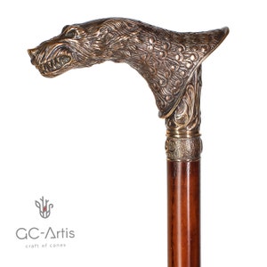 May include: A walking stick featuring a bronze-coloured metal handle shaped like a snarling wolf head with detailed fur texture. The handle sits atop a smooth, dark brown wooden shaft, connected by a textured metal collar. The logo GC-Artis is visible.