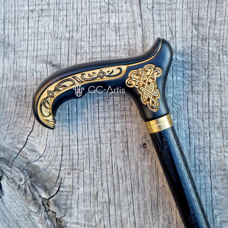 Close-up photo of the GC-Artis Celtic walking cane placed on weathered wood. The handle shows detailed gold engraving, smooth curves and a warm polished finish. The black shaft adds contrast and gives the cane a refined, classic look. The outdoor light highlights the texture of the carving and the natural tones of the wood, showing the cane as a functional yet artistic accessory.