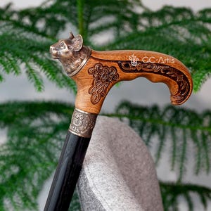 May include: A wooden walking stick with a carved handle featuring a wolf head and intricate Celtic knot designs. The handle is made of light brown wood and the shaft is black.