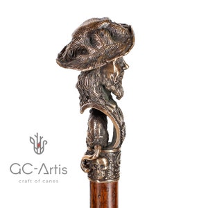 May include: A bronze walking stick with a detailed sculpture of a man's head wearing a hat. The sculpture is attached to a wooden shaft with a skull and crossbones design.