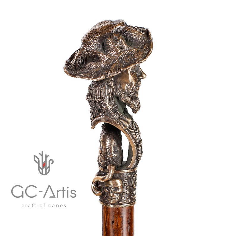 May include: A bronze walking stick with a detailed sculpture of a man's head wearing a hat. The sculpture is attached to a wooden shaft with a skull and crossbones design.