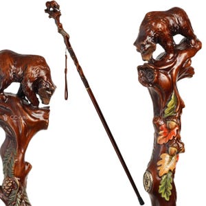 May include: A brown wooden walking stick featuring a hand-carved bear figure at the top. The handle includes detailed carvings of oak leaves and acorns in green, orange, and yellow. A thin brown wrist strap is attached to the shaft, which has a smooth finish and a black rubber tip at the base. The text GC-Artis is visible on the handle.