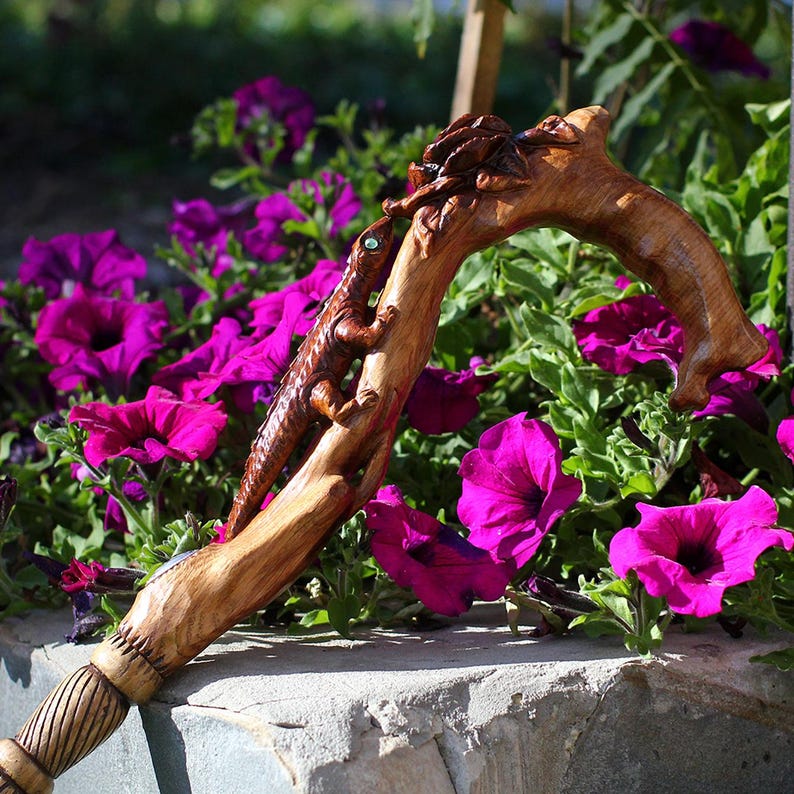 Outdoor photo of the Floral Lizard cane standing near blooming pink flowers. The carved lizard curves around the handle, meeting bright floral elements that catch sunlight. The mix of natural blossoms and the artist’s carved counterparts creates a striking thematic link, showing the cane as decorative, expressive and rooted in nature.