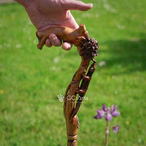 Outdoor photo of a hand holding the Floral Lizard cane near green grass and a small purple flower. The carved lizard shape fits comfortably into the grip, while the carved floral details follow the curve of the handle. Natural lighting brings out warm colors and the expressive handcrafted theme.