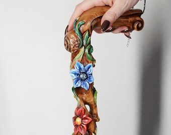 Puede incluir: Un bastón de madera con un diseño floral tallado. El bastón presenta un mango con un diseño de voluta y está adornado con flores azules y rojas, y enredaderas verdes. El bastón es sostenido por una persona.