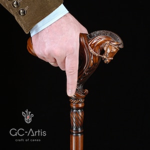 May include: A dark brown wooden walking cane featuring a detailed hand-carved horse head handle with a bridle and mane. The shaft has a smooth, polished finish. The logo GC-Artis craft of canes is visible in the lower left corner of the image.