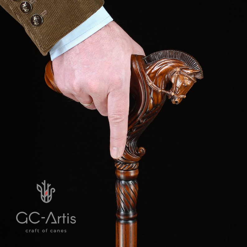 May include: A dark brown wooden walking cane featuring a detailed hand-carved horse head handle with a bridle and mane. The shaft has a smooth, polished finish. The logo GC-Artis craft of canes is visible in the lower left corner of the image.