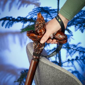 May include: A wooden walking stick with a carved wolf head handle. The handle is dark brown and the stick is a lighter brown. The stick has a black strap with the text "GC-Artis craft of hands" on it.