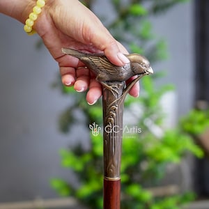 May include: A bronze walking stick with a detailed bird sculpture on top. The bird is perched on a branch and has its beak open as if it is singing. The stick is made of wood and has a brown finish.