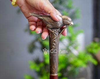 May include: A bronze walking stick with a detailed bird sculpture on top. The bird is perched on a branch and has its beak open as if it is singing. The stick is made of wood and has a brown finish.