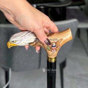May include: A walking cane featuring a carved wooden handle shaped like a bald eagle head with a yellow beak. The handle includes a relief of the Great Seal of the United States with the text IN GOD WE TRUST and a black shaft with a brass collar.