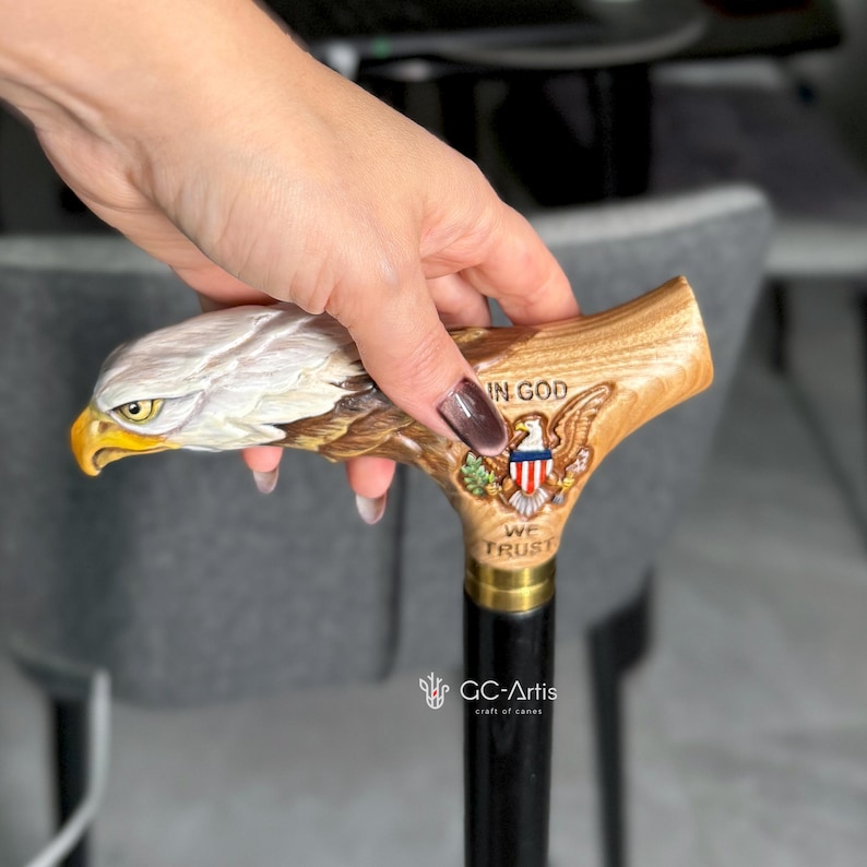 May include: A walking cane featuring a carved wooden handle shaped like a bald eagle head with a yellow beak. The handle includes a relief of the Great Seal of the United States with the text IN GOD WE TRUST and a black shaft with a brass collar.