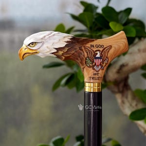 May include: A wooden walking cane handle carved into the shape of a bald eagle head with a yellow beak. The side features an engraved eagle emblem with the text IN GOD WE TRUST. A gold-colored metal collar connects the handle to a black shaft.