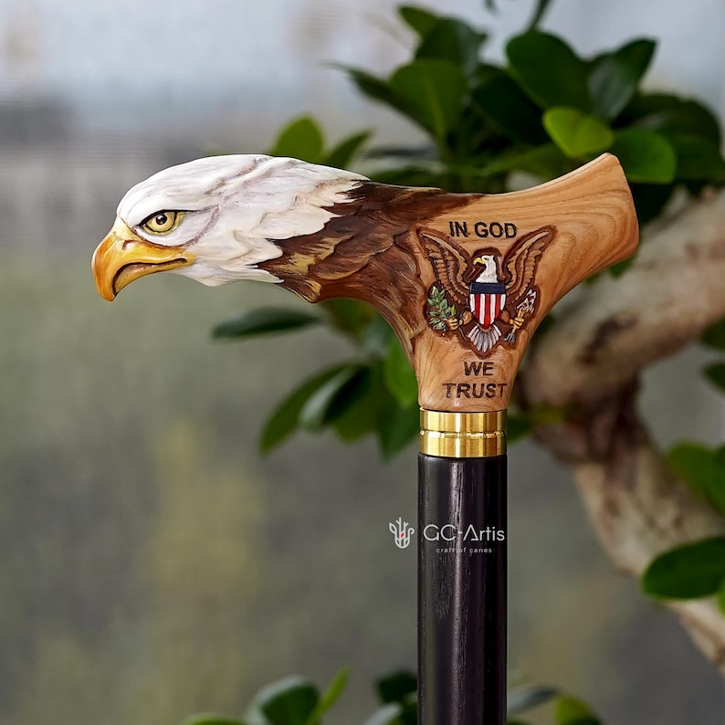 May include: A wooden walking cane handle carved into the shape of a bald eagle head with a yellow beak. The side features an engraved eagle emblem with the text IN GOD WE TRUST. A gold-colored metal collar connects the handle to a black shaft.