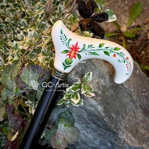 Puede incluir: Un bast&oacute;n blanco con un mango negro. El mango est&aacute; decorado con un dise&ntilde;o floral pintado a mano con colores rojo, verde y naranja. El texto "GC-Artis craft of canes" es visible en el mango.