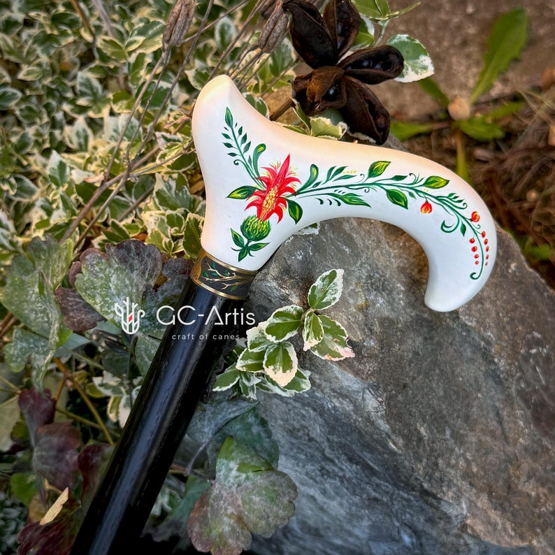 Puede incluir: Un bast&oacute;n blanco con un mango negro. El mango est&aacute; decorado con un dise&ntilde;o floral pintado a mano con colores rojo, verde y naranja. El texto "GC-Artis craft of canes" es visible en el mango.