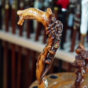 Close-up outdoor photo of the cane against a wooden railing with bright flowers behind it. The carved lizard wraps down the handle with textured scales and flowing shapes. The floral carvings add colorful accents. The warm wood finish and organic contours give the cane an artistic, handcrafted presence in a lively outdoor setting.
