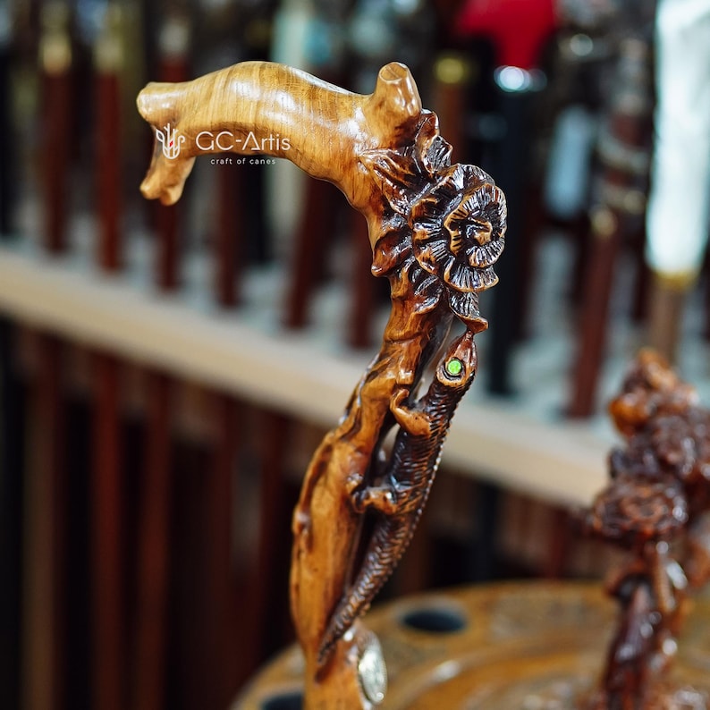 Close-up outdoor photo of the cane against a wooden railing with bright flowers behind it. The carved lizard wraps down the handle with textured scales and flowing shapes. The floral carvings add colorful accents. The warm wood finish and organic contours give the cane an artistic, handcrafted presence in a lively outdoor setting.