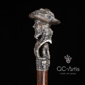 May include: A silver walking stick with a detailed sculpted head of a pirate wearing a hat. The stick has a wooden shaft and a decorative skull design at the bottom.