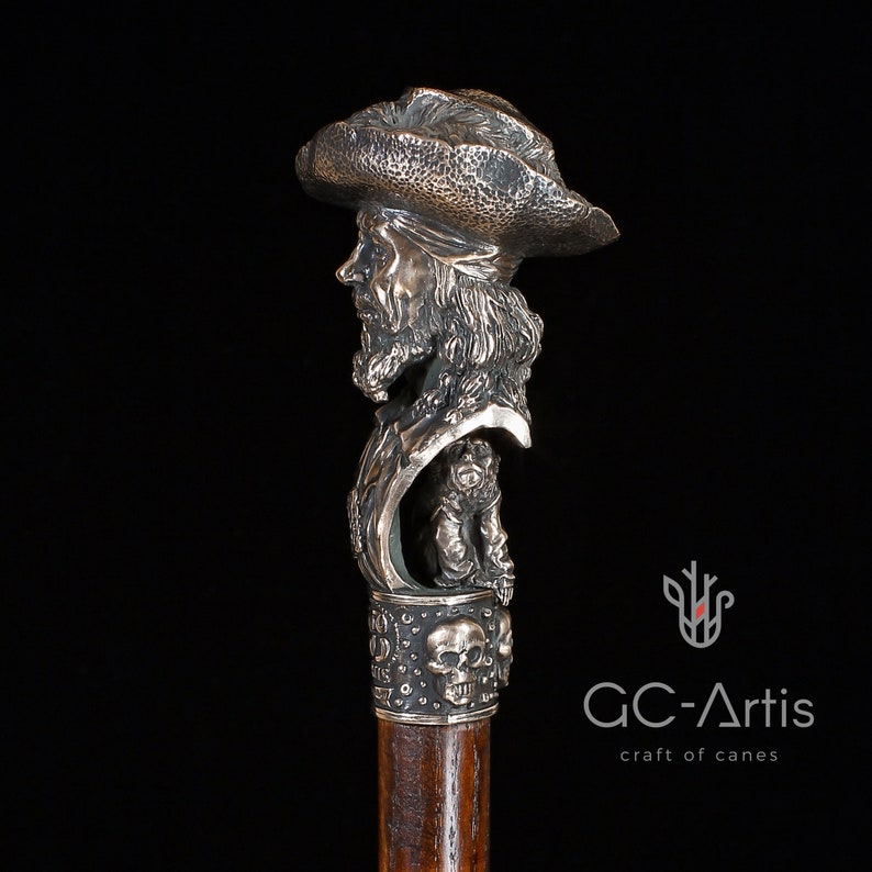 May include: A silver walking stick with a detailed sculpted head of a pirate wearing a hat. The stick has a wooden shaft and a decorative skull design at the bottom.
