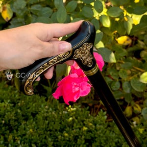 Outdoor photo of the Celtic cane held near a blooming pink rose. The engraved handle with gold accents contrasts with the bright flower, emphasizing craftsmanship and decorative detail. The black shaft frames the scene, showing how the cane looks in the hand during real outdoor use.