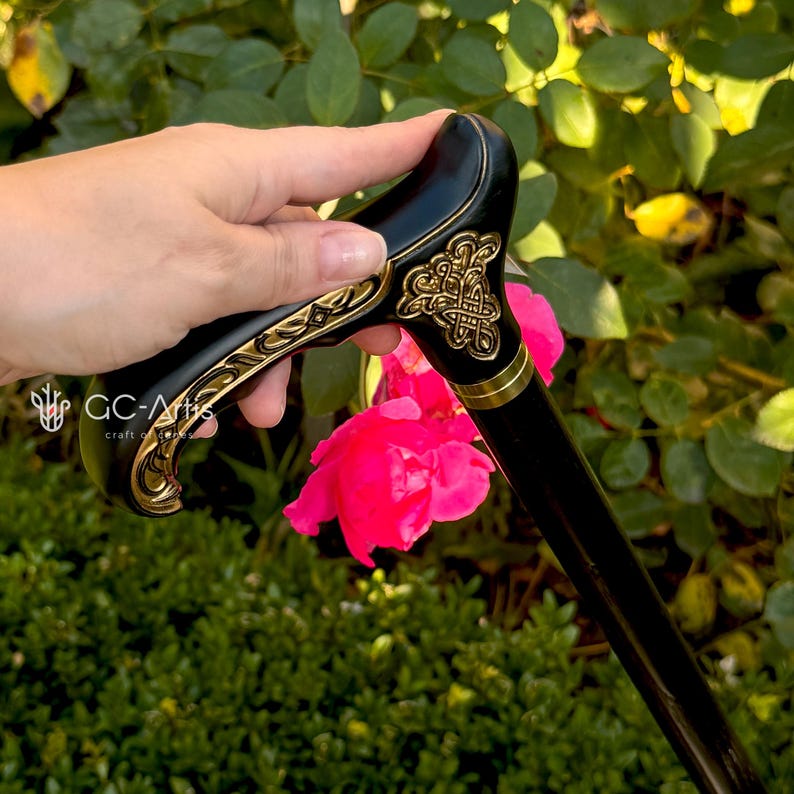Outdoor photo of the Celtic cane held near a blooming pink rose. The engraved handle with gold accents contrasts with the bright flower, emphasizing craftsmanship and decorative detail. The black shaft frames the scene, showing how the cane looks in the hand during real outdoor use.