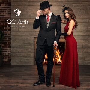 May include: A man in a black tuxedo and a top hat stands next to a woman in a red dress. They are both holding a walking stick. The man is holding the stick in his right hand and the woman is holding the stick in her left hand. The man is looking at the camera and the woman is looking at the man. The background is a brick wall with a fireplace.