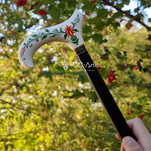 Puede incluir: Un bast&oacute;n blanco con un mango negro. La parte superior del bast&oacute;n est&aacute; pintada con un dise&ntilde;o floral con flores rojas y hojas verdes. El texto "GC-Artis craft of canes" es visible en el bast&oacute;n.