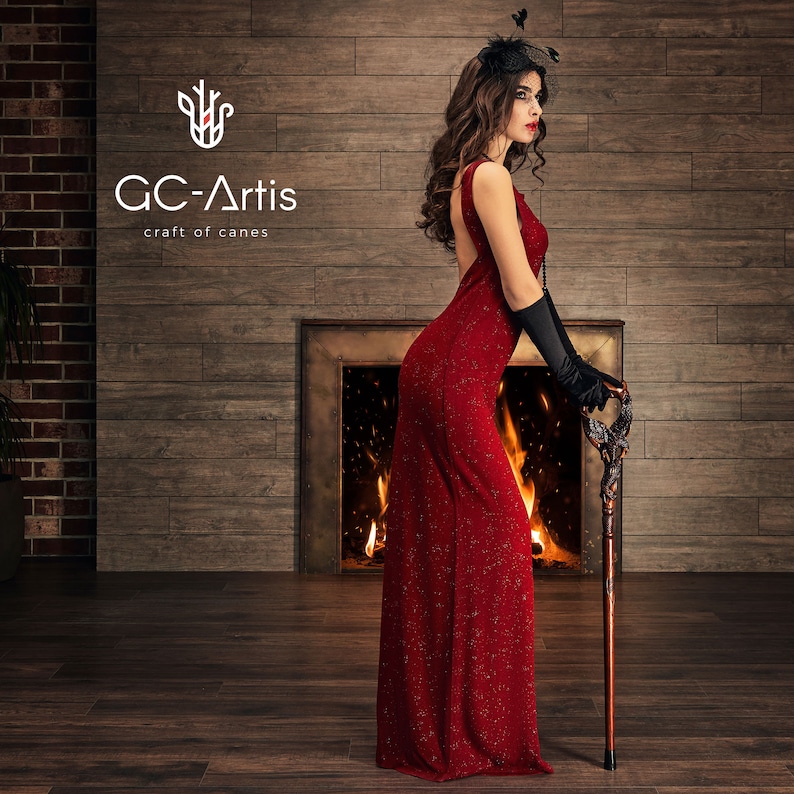 May include: A woman in a red dress and black gloves stands in front of a fireplace holding a wooden walking stick with a carved bird design. The text "GC-Artis craft of canes" is visible in the upper left corner of the image.