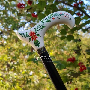 Puede incluir: Un bast&oacute;n blanco con un dise&ntilde;o floral pintado a mano con flores rojas, verdes y blancas. El bast&oacute;n tiene un eje negro con una banda dorada y el texto "GC-Artis craft of canes".