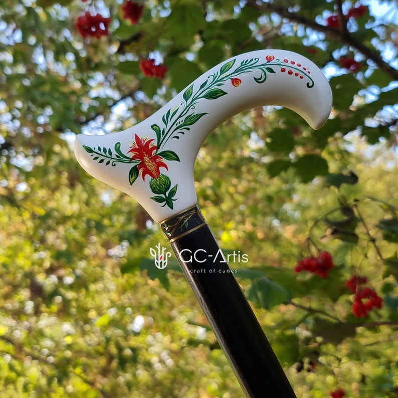 Puede incluir: Un bast&oacute;n blanco con un dise&ntilde;o floral pintado a mano con flores rojas, verdes y blancas. El bast&oacute;n tiene un eje negro con una banda dorada y el texto "GC-Artis craft of canes".