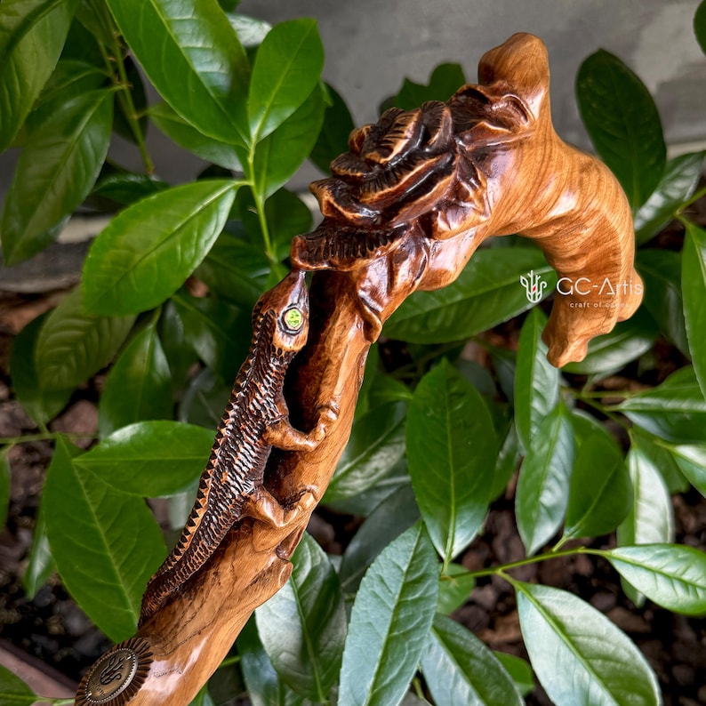Photo of the Floral Lizard cane resting among glossy green leaves. The carved lizard and floral motifs wrap around the handle and upper shaft, painted with subtle color accents. Natural outdoor lighting enhances sculpted details and the warm honey-brown wood finish, giving the cane a lively, organic feel inspired by forest and garden themes.