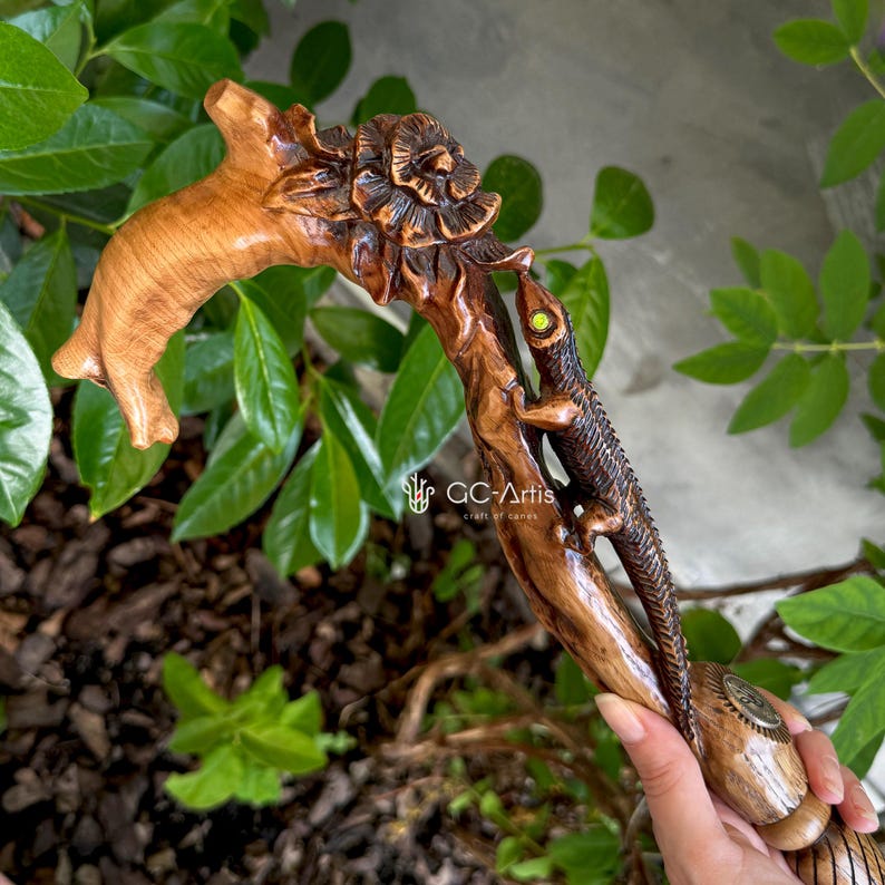 Close-up photo of the GC-Artis Floral Lizard walking cane held outdoors among bright green leaves. The carved handle combines a stylized lizard shape with raised floral elements, blending warm wood tones with hand-painted accents. The texture of the carving stands out in natural light, showing detailed scales, petals and curved lines that give the cane a dynamic, nature-inspired personality.
