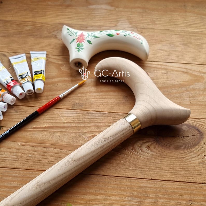 Puede incluir: Un bast&oacute;n de madera con un acabado de madera natural y una banda de lat&oacute;n. El bast&oacute;n tiene un mango curvo y se est&aacute; pintando con pinturas acr&iacute;licas. El texto "GC-Artis craft of canes" es visible en el bast&oacute;n.
