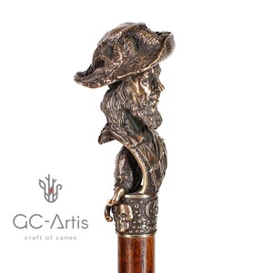 May include: A bronze walking stick with a detailed sculpture of a man's head wearing a hat. The sculpture is attached to a wooden shaft with a skull and crossbones design.
