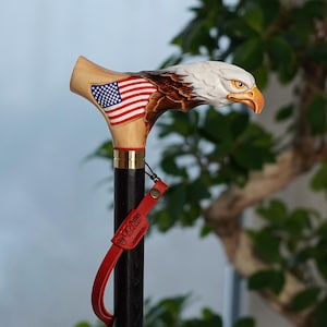 以下が含まれることがあります： アメリカ国旗が側面に描かれた、木彫りの鷲の頭部を模したハンドル付きの杖。鷲は白い羽、茶色のアクセント、黄色い嘴が特徴。金色の金属製カラーがハンドルと黒いシャフトを繋いでいる。赤いレザーストラップには「GC-Artis」の刻印がある。