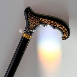 Studio photo of the Celtic cane near a soft rainbow reflection on a white wall. The engraved handle appears warm and detailed, while the black shaft keeps the silhouette simple. The subtle color reflection adds a gentle, artistic touch to the scene, highlighting elegance and craftsmanship.