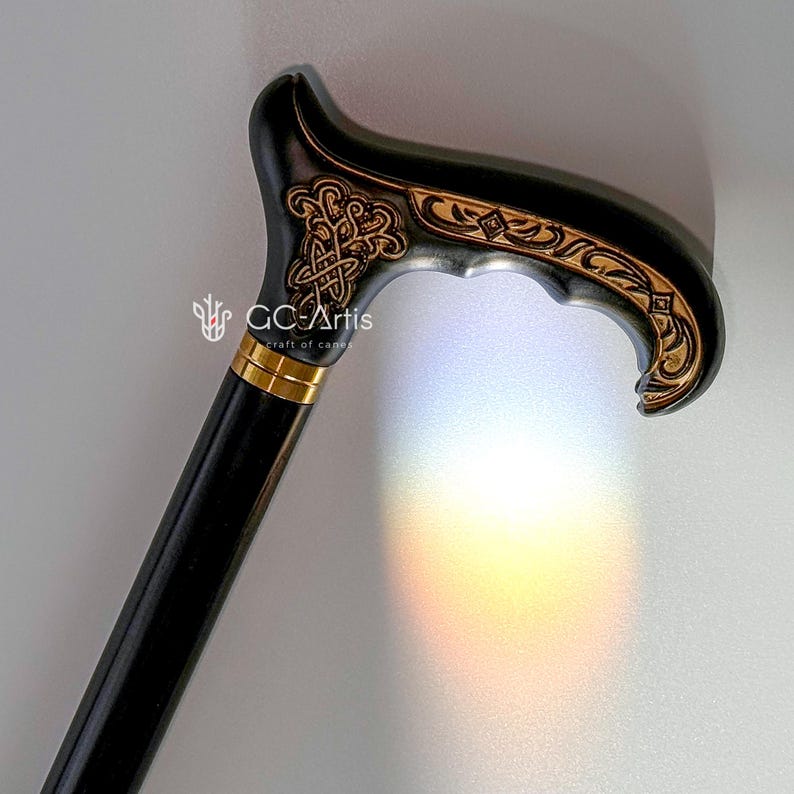 Studio photo of the Celtic cane near a soft rainbow reflection on a white wall. The engraved handle appears warm and detailed, while the black shaft keeps the silhouette simple. The subtle color reflection adds a gentle, artistic touch to the scene, highlighting elegance and craftsmanship.