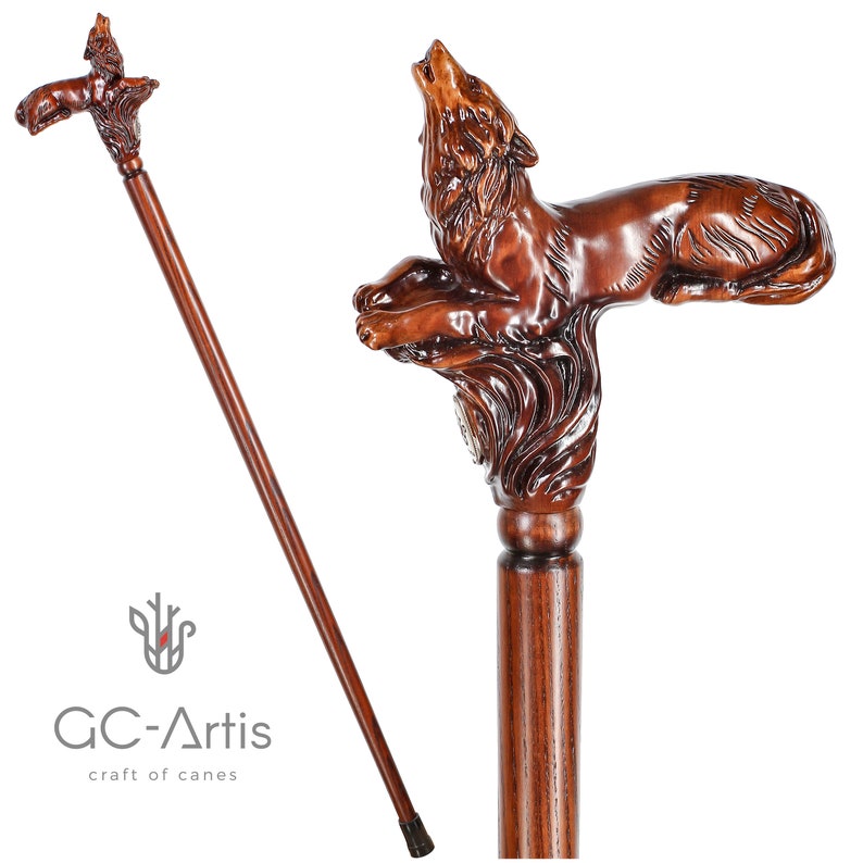 May include: A wooden walking stick with a carved wolf head handle. The wolf is howling and has a detailed, realistic design. The stick is brown and has a dark brown finish. The text "GC-Artis craft of canes" is visible on the stick.