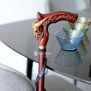 May include: A wooden walking stick with a detailed skull design and a black handle. The handle is engraved with the text "G.C. Artis" and "Craft of Canes".